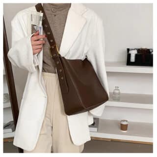 Photo d'une femme portant un sac fourre-tout bandoulière de style vintage marron. La femme porte un pull col roulé marron, une veste blanche et un pantalon beige, elle se tient debout devant des étagères.
