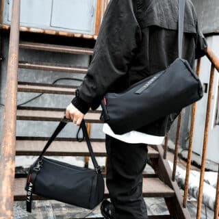 Homme portant à la main et sur l'épaule des sacs de sport à bandoulière multifonction noirs. L'homme est habillé en tenue de sport noire et monte des escalier en acier.