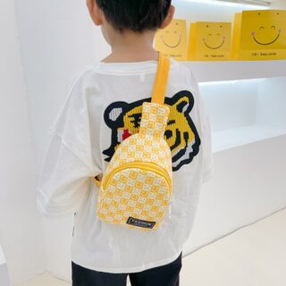 On voit un petit garçon brun de dos qui porte un sac jaune avec des smileys, en bandoulière. Il porte un tee-shirt blanc avec un tigre et un pantalon noir.