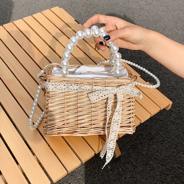 Photo d'un petit sac en osier entouré d'un ruban de dentelle avec une anse et une bandoulière en perles. Le sac est tenue par une main au dessus d'une table en bois