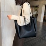 Femme en pull blanc poortant en bandoulière un grand sac noir à l'intérieur d'un batiment