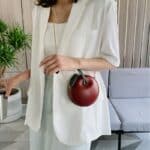 Femme dans une salon avec une plante et un canapé gris portant une veste blanche et un sac rouge en forme de pomme.