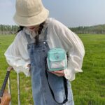 On voit une fille dans un champs qui porte un bob et une robe salopette en jean et un chemisier. Elle porte un sac à bandoulière bleu ciel.