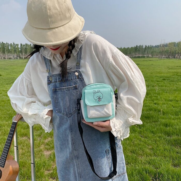 On voit une fille dans un champs qui porte un bob et une robe salopette en jean et un chemisier. Elle porte un sac à bandoulière bleu ciel.