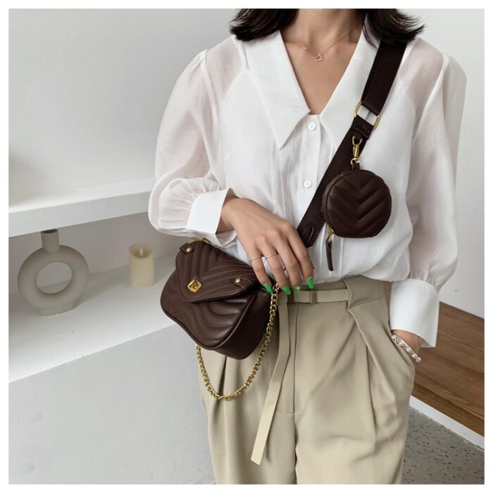 Femme portant un sac en cuir synthétique marron en bandoulière avec une petite pochette ronde sur la sangle bandoulière. Le sac a aussi une chaine. La femme se tient debout devant un mur étagère, elle est habillée avec un pantalon beige et une chemise blanche.
