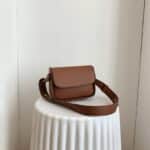 Photo d'un sac pochette bandoulière en cuir synthétique marron de syle minimaliste posé sur un présentoir blanc texturé