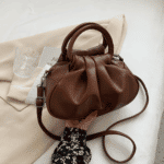 Photo d'un petit sac à main bandoulière marron aux détails plissé tenue par une main au dessus d'une veste blazer beige.