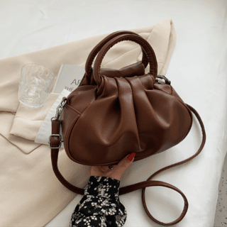 Photo d'un petit sac à main bandoulière marron aux détails plissé tenue par une main au dessus d'une veste blazer beige.